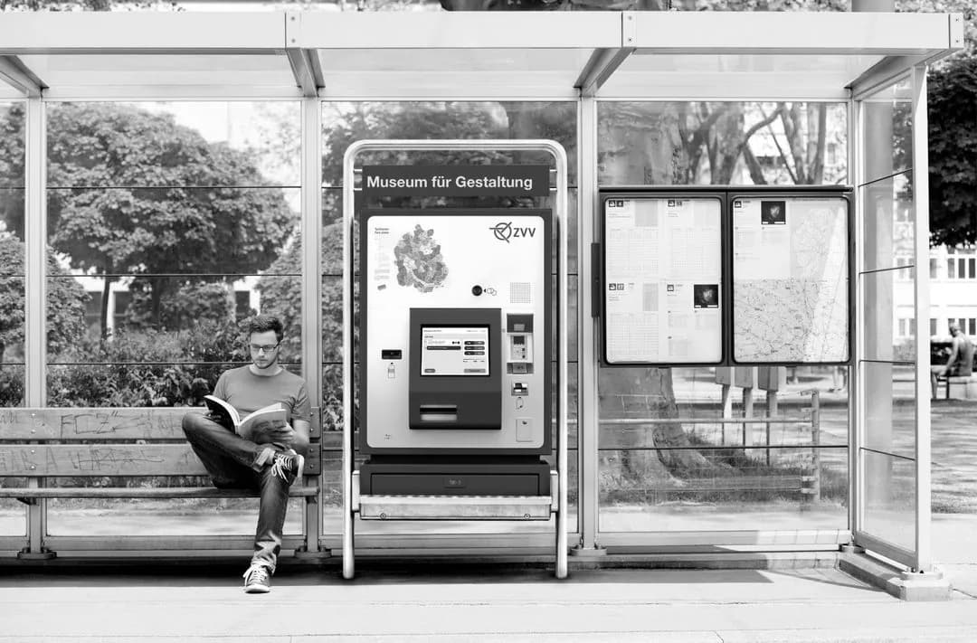Black-and-white photograph of a person reading on a bench beside a poster stand and ticket machine at a bus stop