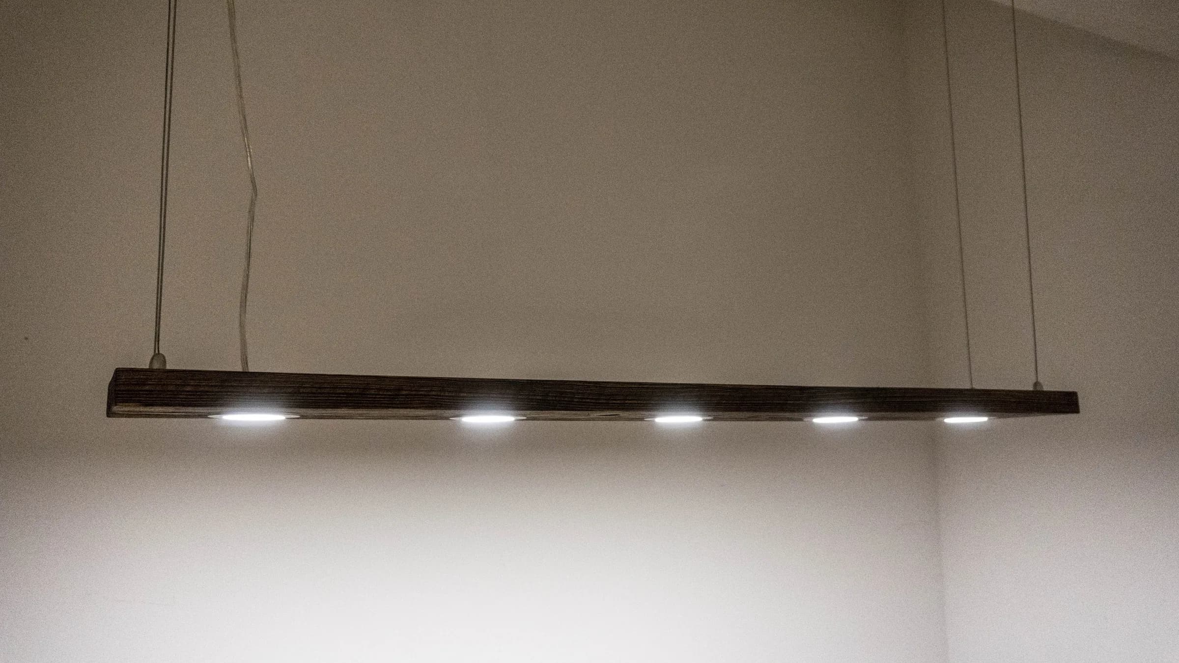 Long black bar light with five spots suspended on thin cables against a white wall